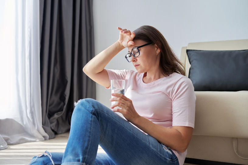 A woman in pain sits on the floor with a glass of water in her hand.