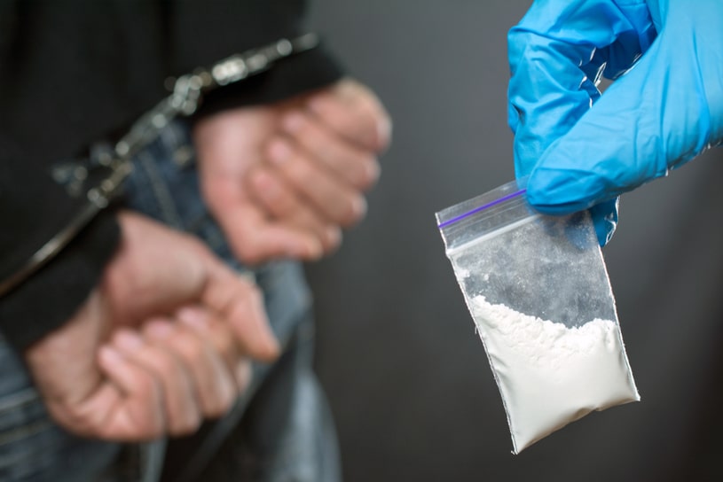 A hand in a glove holds a prohibited substance against the background of a guy in handcuffs.